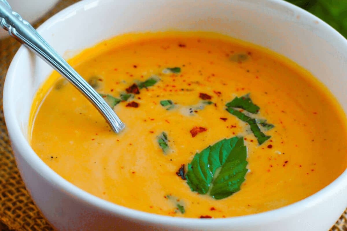 A bowl of creamy orange soup, one of our cozy vegetable soups, garnished with chopped herbs, red pepper flakes, and a sprig of fresh basil, with a spoon resting inside—perfect to warm up your day.