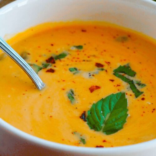 A bowl of creamy orange soup from our cozy vegetable soups collection, garnished with fresh herbs and spices, with a spoon resting inside the bowl—perfect to warm up on a chilly day.