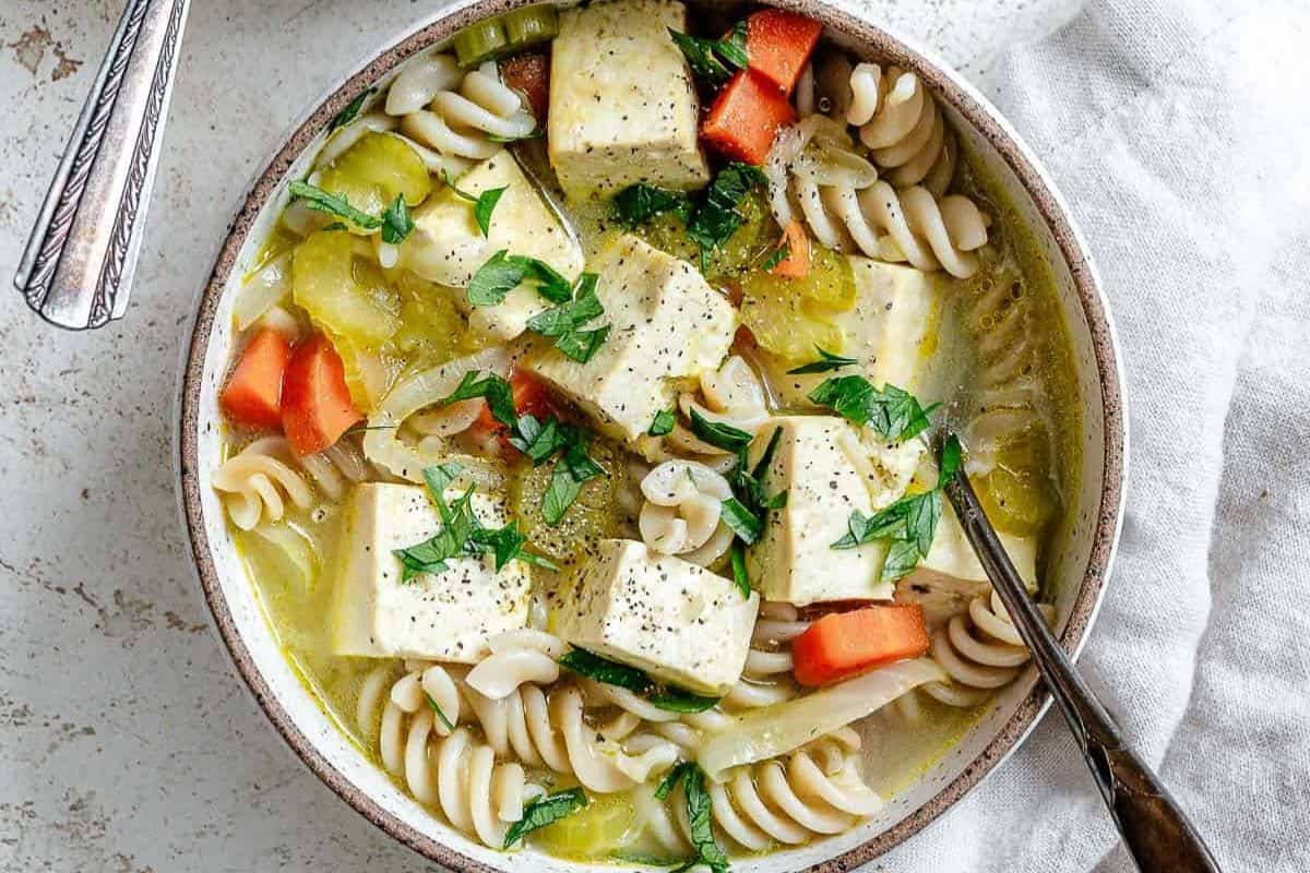 A bowl of comforting soup with rotini pasta, cubed tofu, carrots, celery, and parsley garnish, served with a spoon on a light surface—perfect to warm your soul.