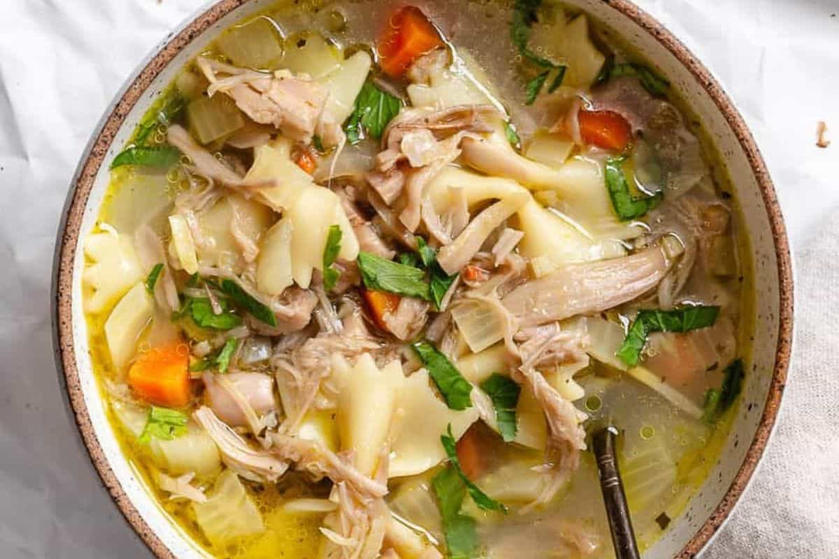Bowl of comforting chicken soup with shredded chicken, carrots, celery, noodles, and herbs, with a spoon inside—one of the most soul warming soups.