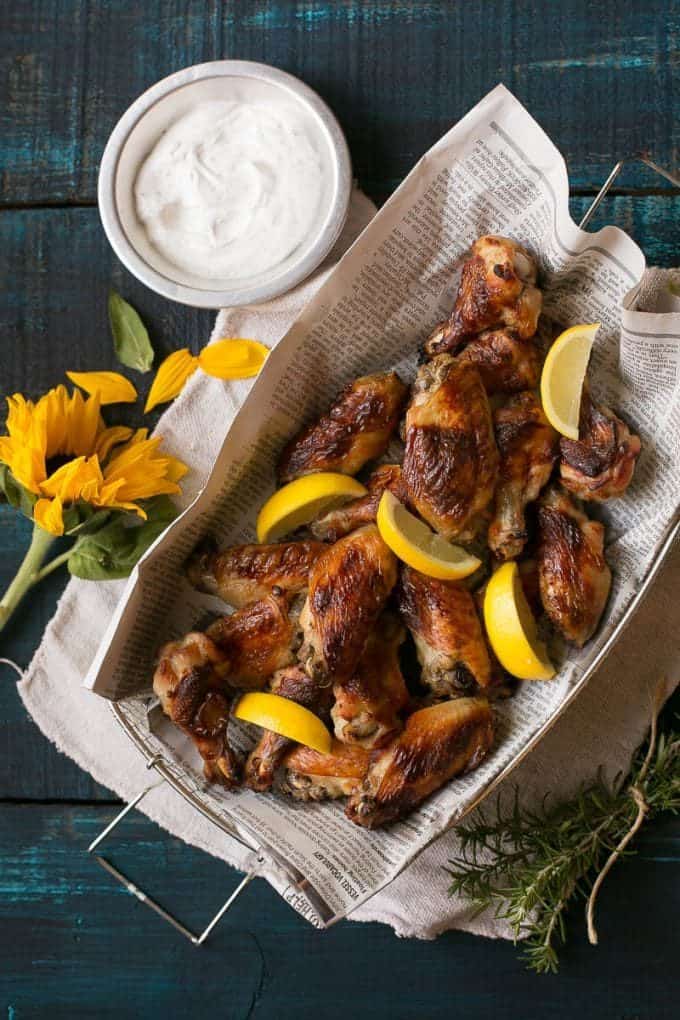 Basket of roasted chicken wings with lemon wedges on newspaper, served with a bowl of white dipping sauce. Sunflower and rosemary are beside the basket on a dark wooden table, making it the perfect scene for trying out new finger-lickin’ wing recipes.