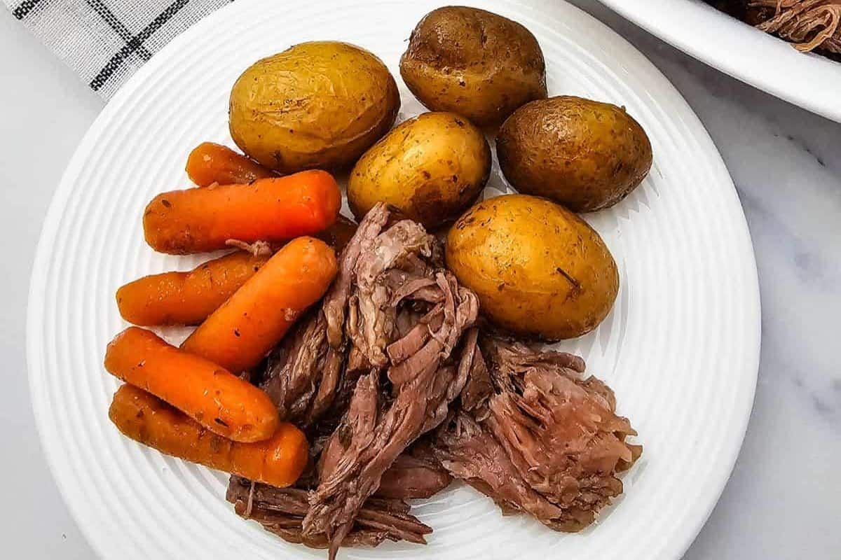 A white plate with cooked baby potatoes, whole carrots, and shredded pot roast arranged in separate sections—this family friendly meal is inspired by chuck roast recipes everyone loves.