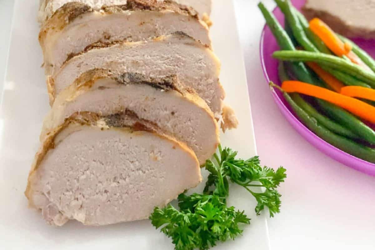 Sliced roasted pork loin arranged on a white plate, garnished with parsley, with a side of green beans and carrots—this family-friendly meal is sure to be one everyone loves.