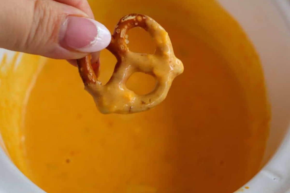A hand holds a pretzel dipped in orange cheese sauce above a bowl filled with the same sauce—perfect for enjoying your favorite Slow Cooker Dips.