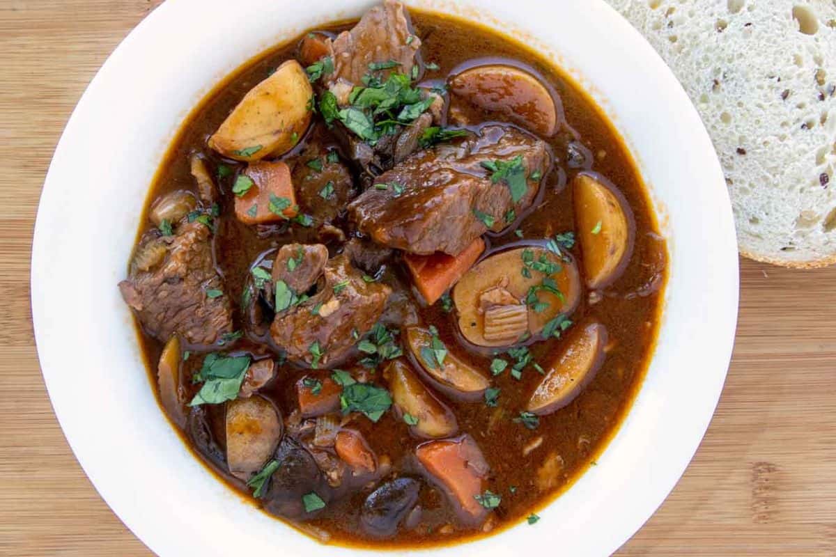 A bowl of beef stew made with tender chuck roast, potatoes, carrots, and herbs in a brown broth, served with a slice of bread on the side—this family friendly meal is one everyone loves.