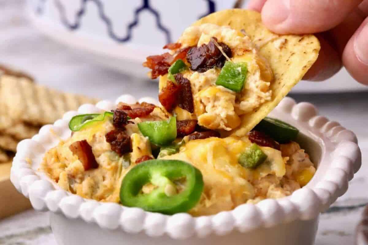 A hand dips a tortilla chip into a bowl of creamy cheese dip topped with bacon bits and sliced jalapeños—one of the ultimate Slow Cooker Dips for any gathering.