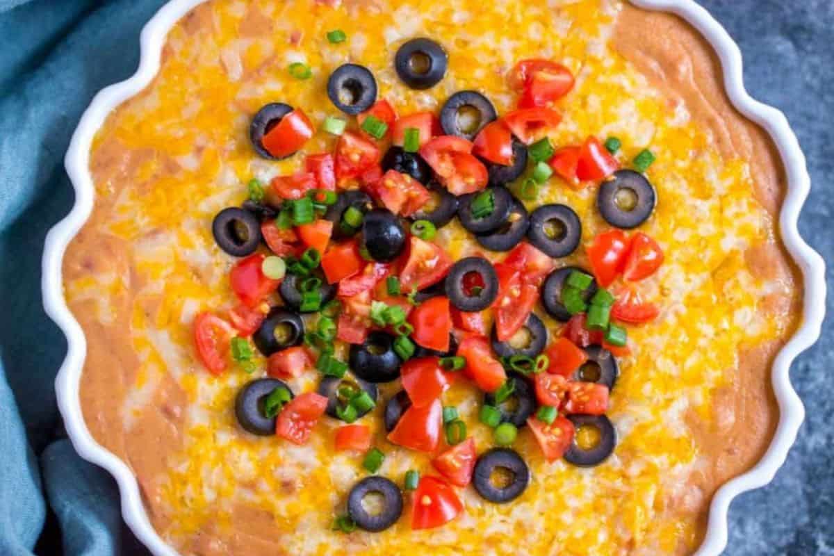 A round dish of layered dip, perfect for Slow Cooker Dips, topped with shredded cheese, sliced black olives, chopped tomatoes, and green onions.