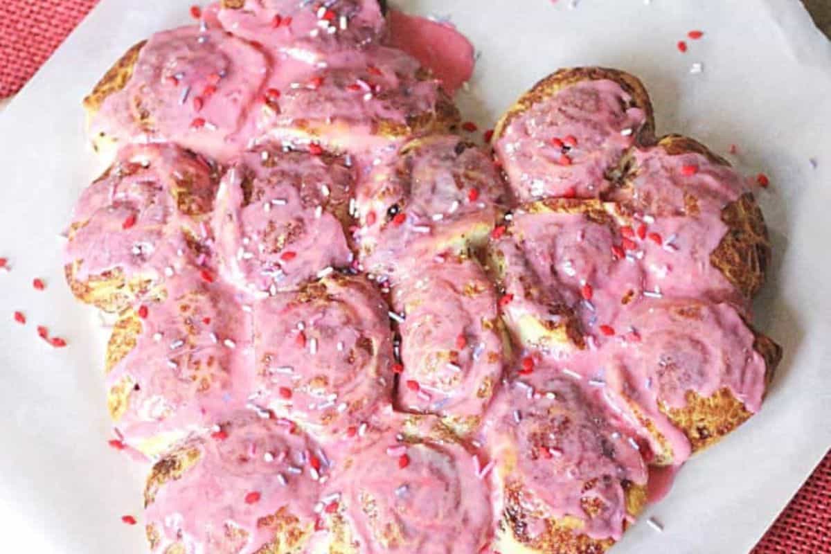 Heart-shaped cinnamon rolls topped with pink icing and red and white sprinkles, arranged on parchment paper—a perfect Valentine's Day breakfast to sweeten your brunch recipes.