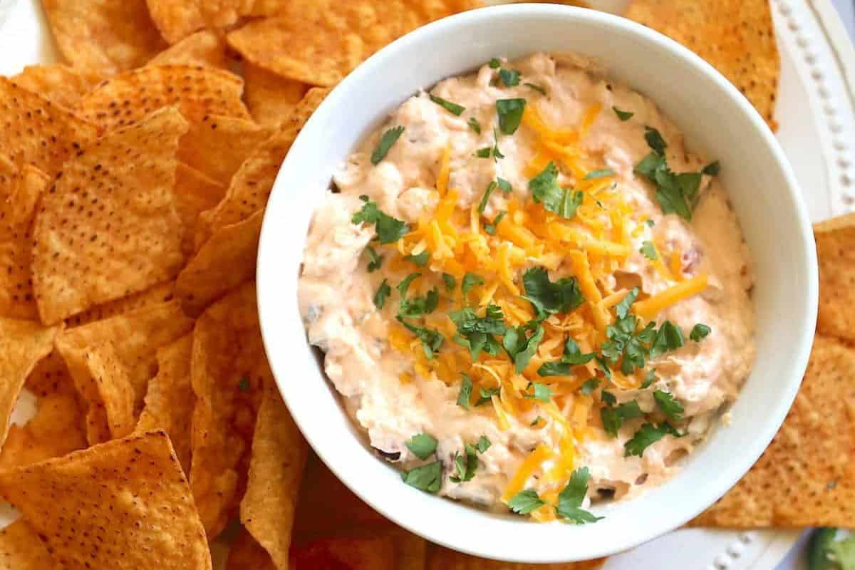 A bowl of creamy Slow Cooker Dip topped with shredded cheese and herbs is surrounded by triangular tortilla chips on a white plate.