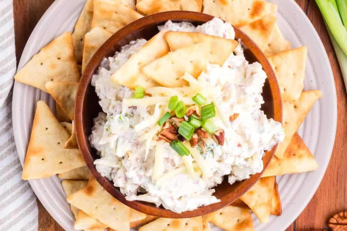 A bowl of creamy Slow Cooker Dip topped with shredded cheese, green onions, and pecans, surrounded by triangular crackers on a plate.