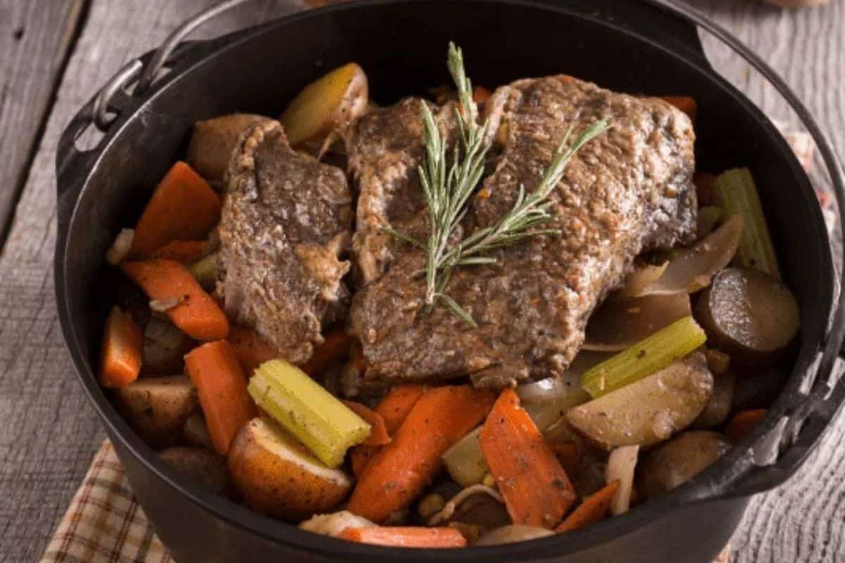 A cast iron pot filled with tender pot roast, potatoes, carrots, celery, and onions—one of those Chuck Roast Recipes that’s truly family friendly—garnished with a sprig of rosemary.