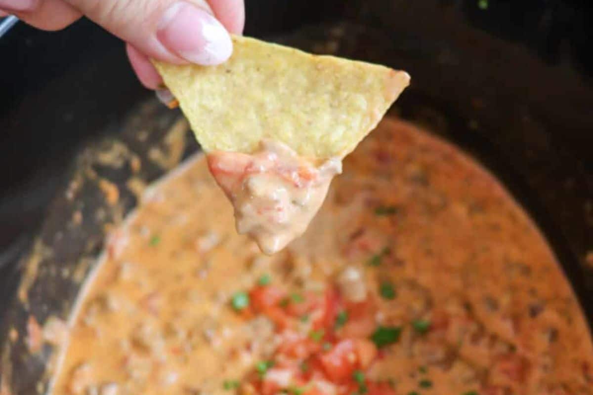 A hand holding a tortilla chip dipped in creamy cheese dip with diced tomatoes and herbs, perfect for enjoying Slow Cooker Dips at your next gathering.