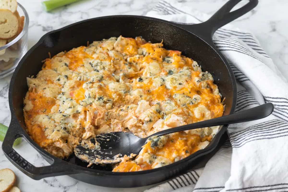 A cast iron skillet filled with baked cheesy dip, partially scooped out with a black spoon, sits on a striped towel atop a marble surface—perfect inspiration for your next round of Slow Cooker Dips.