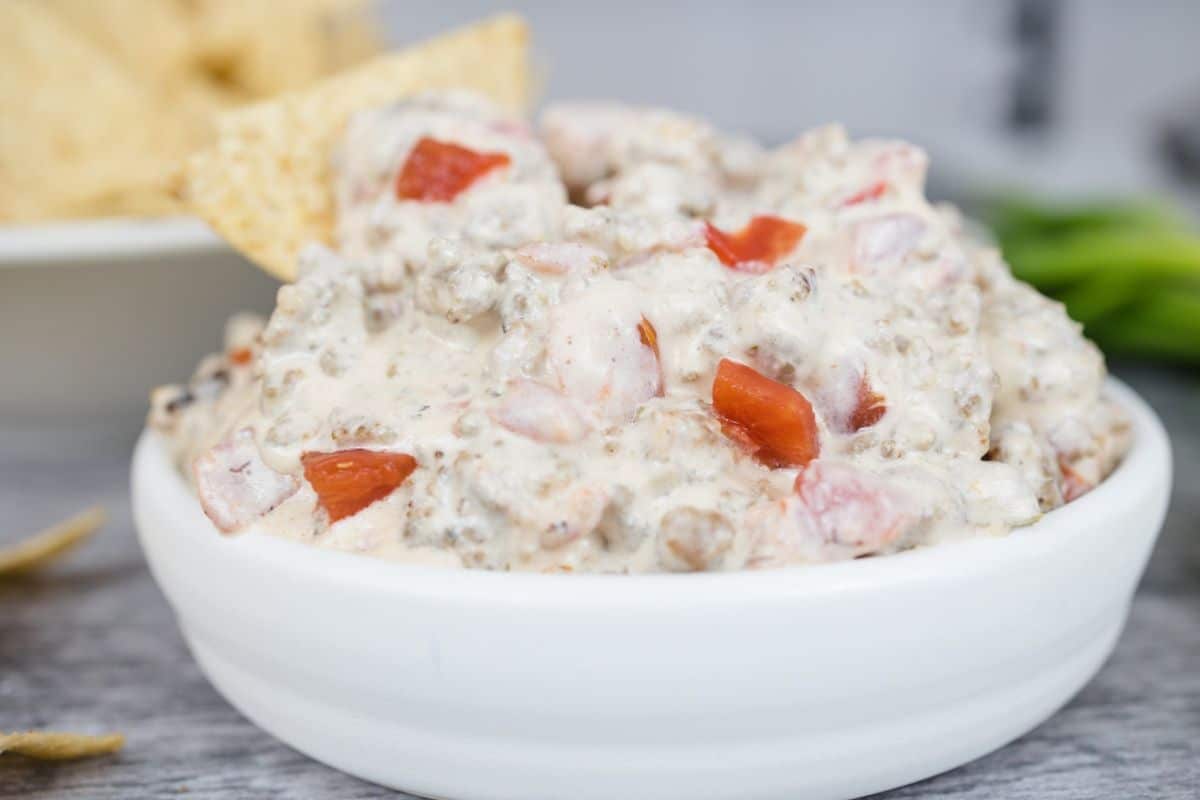 A white bowl filled with creamy sausage dip made from ground meat and diced tomatoes, with a tortilla chip dipped in the side—a perfect choice for easy Slow Cooker Dips at any gathering.