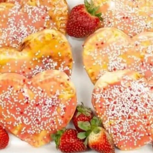 Heart-shaped pastries with pink glaze and white sprinkles, perfect for Galentine’s Day celebrations, are surrounded by fresh strawberries on a white dish.