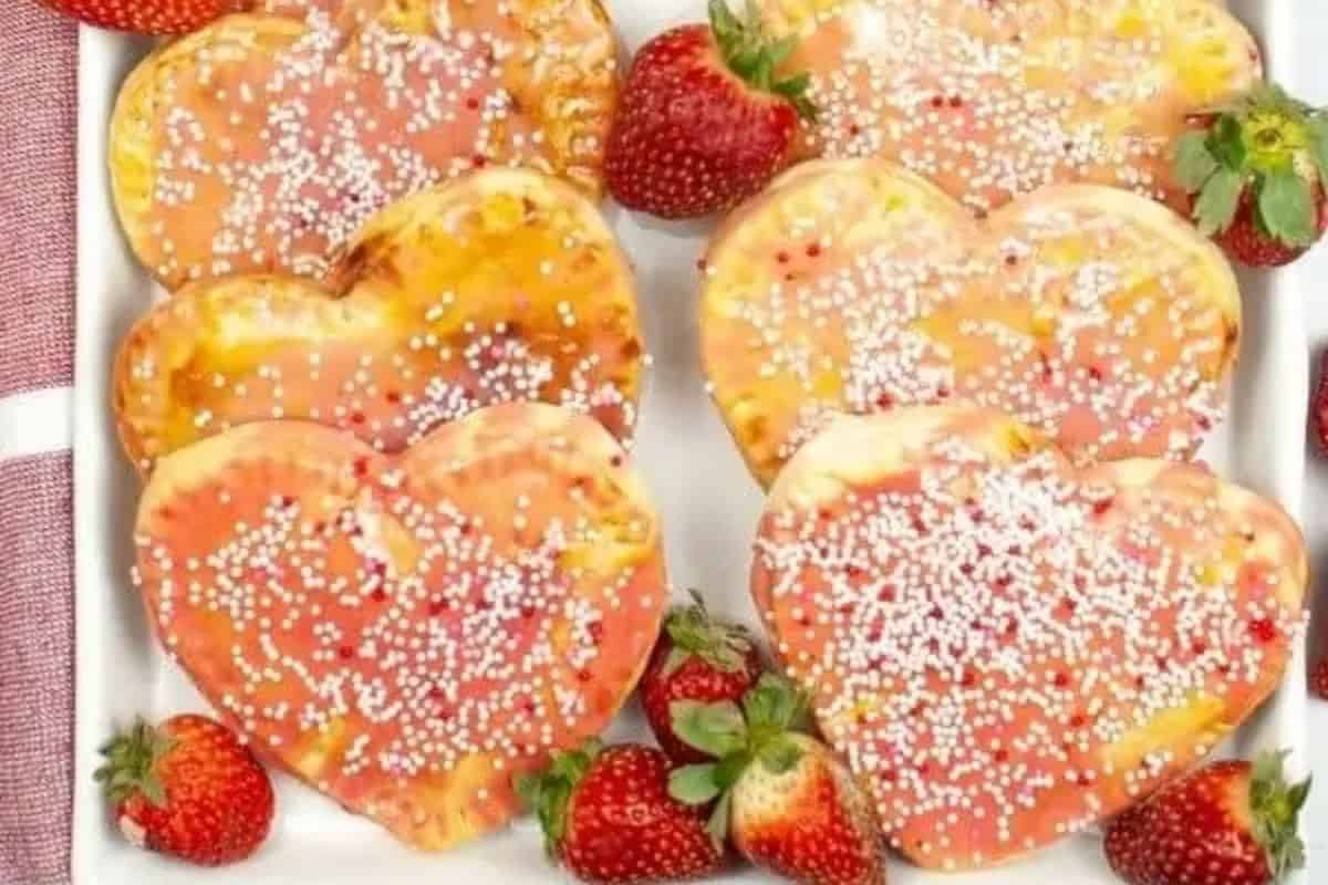 Heart-shaped homemade poptarts with pink glaze and white sprinkles, perfect for Galentine’s Day celebrations, are surrounded by fresh strawberries on a white dish.