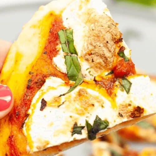 A hand holding a slice of pizza topped with tomato sauce, mozzarella, basil, and a piece of mushroom—a perfect night in comfort and an inspiration for new pizza recipes.