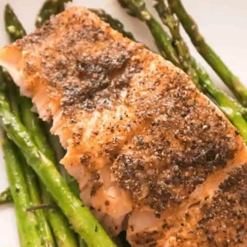 Irresistible Salmon fillet seasoned with pepper, served on a bed of cooked asparagus on a white plate.