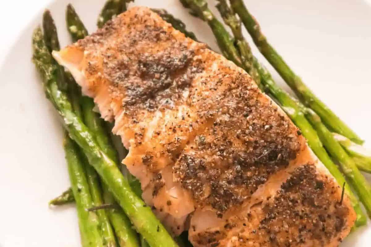 Irresistible Salmon fillet seasoned with pepper, served on a bed of cooked asparagus on a white plate.