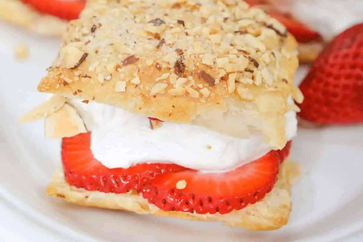 A festive delight for Galentine’s Day, this amaretto strawberry pastry features sliced strawberries and whipped cream nestled between flaky, nut-topped pastry layers on a white plate. Perfect for celebrating with your bestie!.