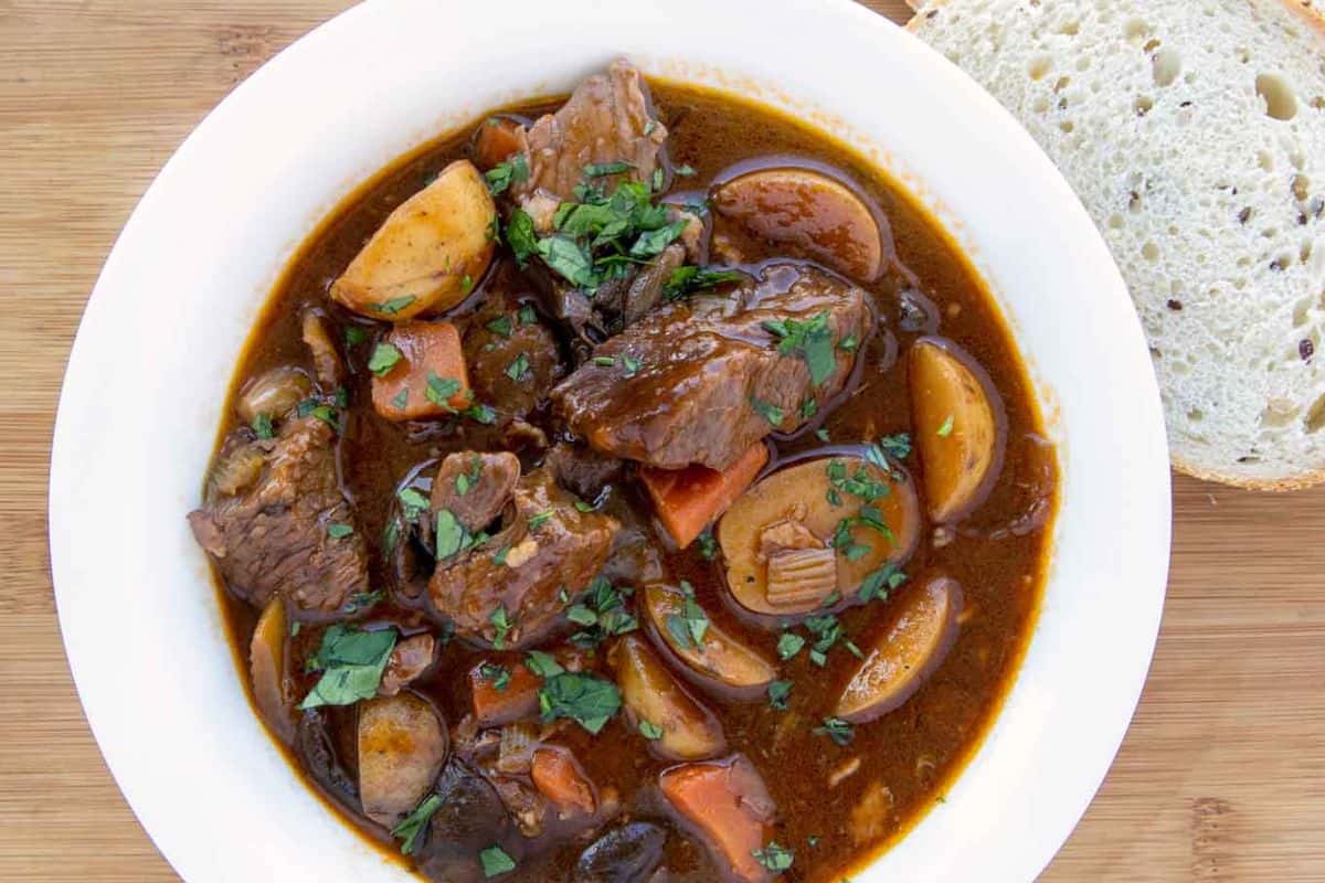 A bowl of Irish-inspired beef stew with carrots, potatoes, and herbs, alongside a slice of bread on a wooden surface—comfort food to warm your soul.