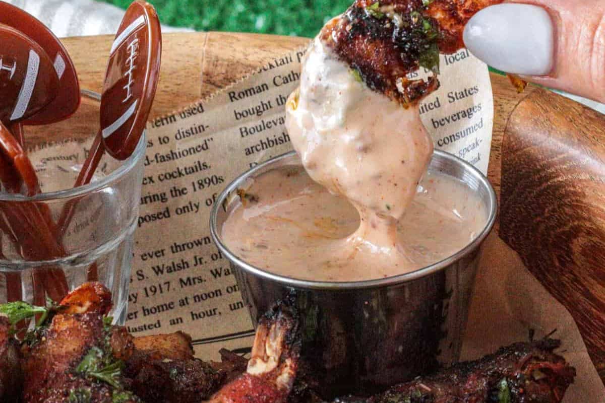 Bacon wrapped shrimp dipped in sauce, held above a metal cup, showcasing home cooking with restaurant quality. Football-themed toothpicks are in the background.