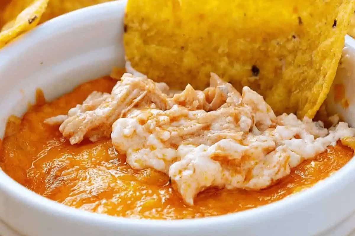 This crowd-pleasing buffalo chicken dip, adorned with a shredded topping in a white bowl, is perfect for any potluck gathering. Serve it with a tortilla chip for an irresistible treat.