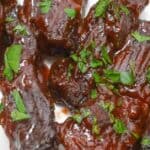 Baked Country Style Ribs coated in a thick, dark barbecue sauce and garnished with chopped fresh parsley are served on a white plate.
