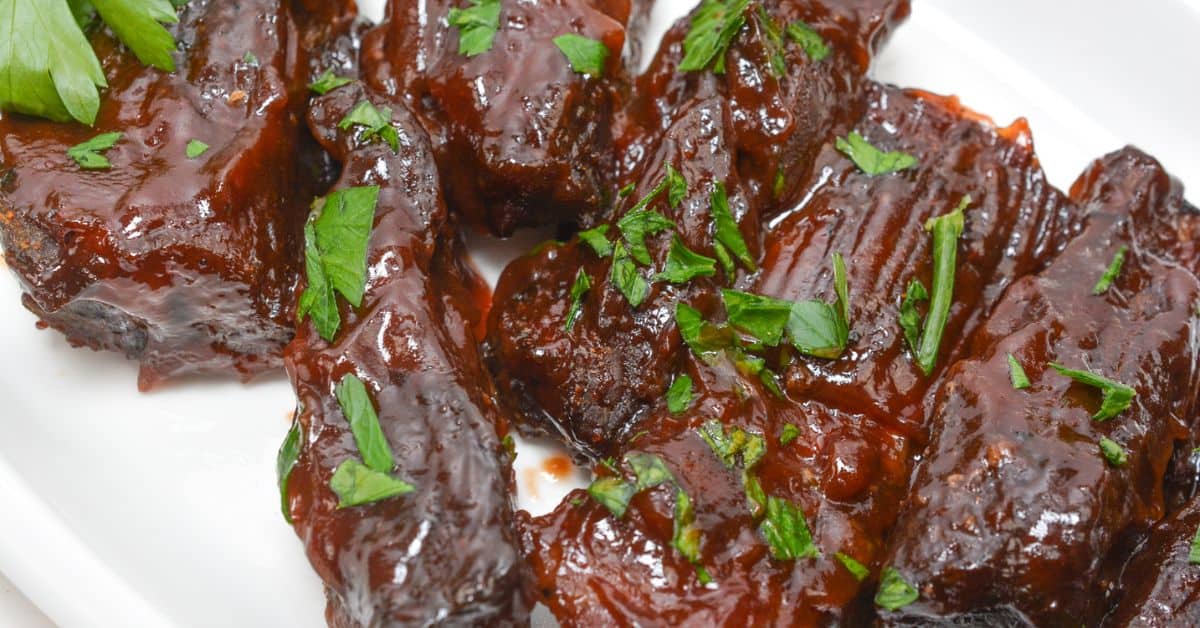 Baked Country Style Ribs coated in a thick, dark barbecue sauce and garnished with chopped fresh parsley are served on a white plate.