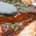 A pair of tongs holds glazed meat garnished with chopped herbs, with more blurred in the background.