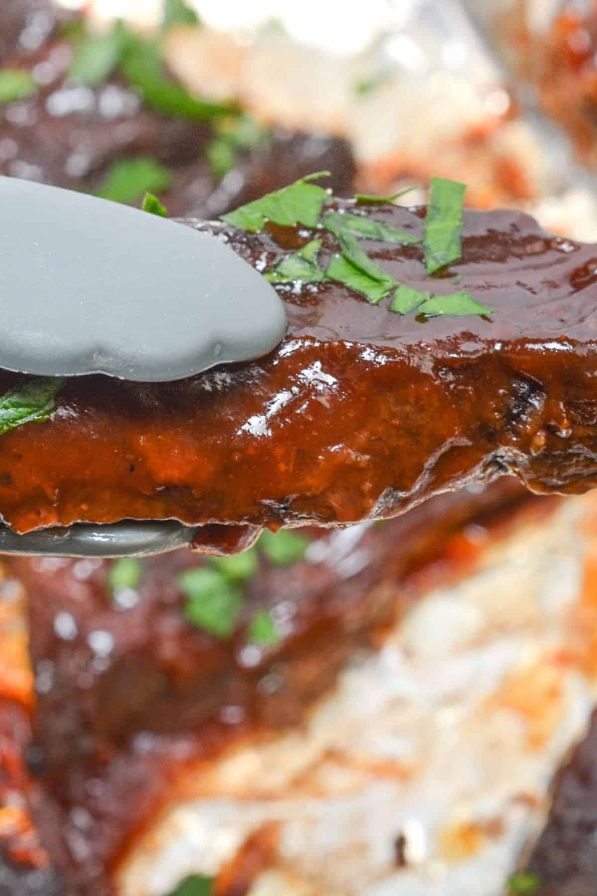 A pair of tongs holds glazed meat garnished with chopped herbs, with more blurred in the background.