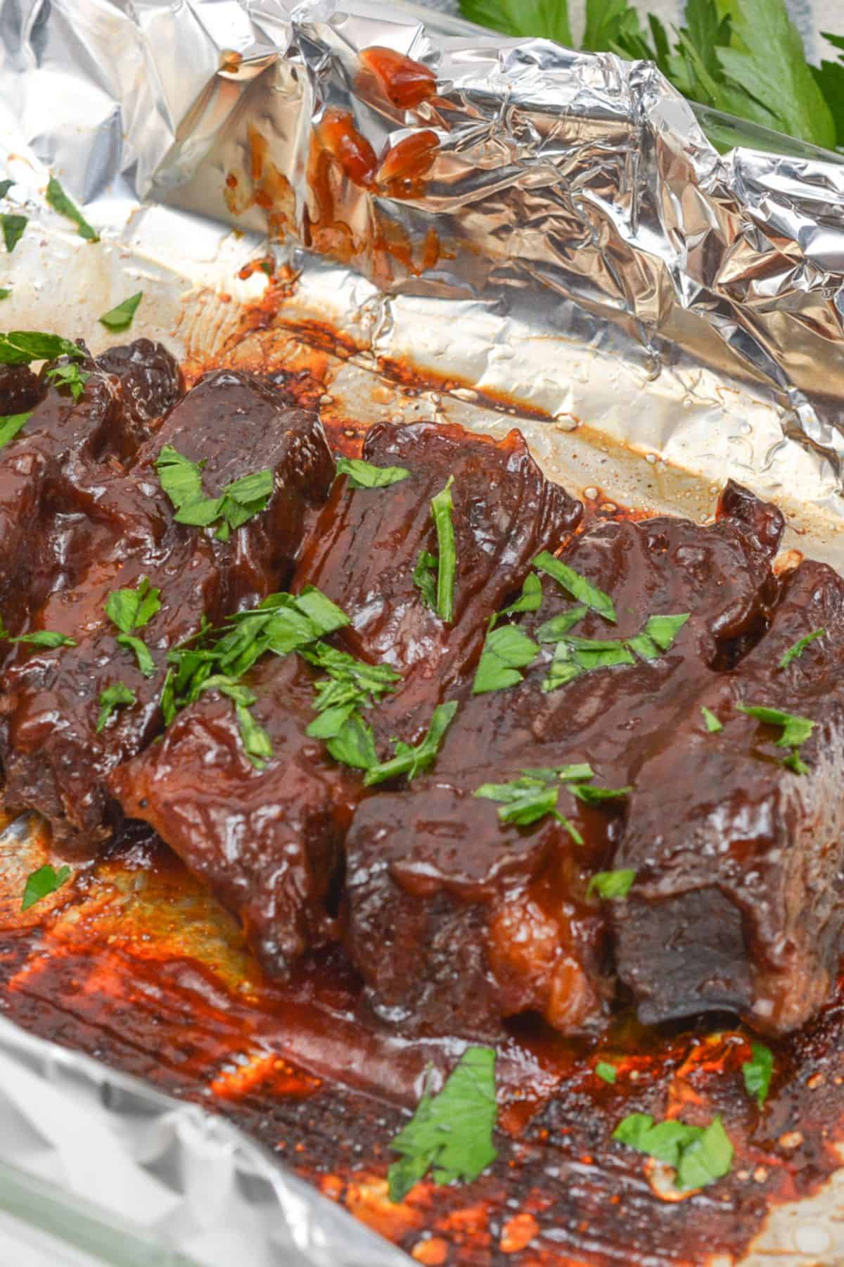 Several pieces of glazed, cooked meat garnished with chopped parsley on a foil-lined baking tray.