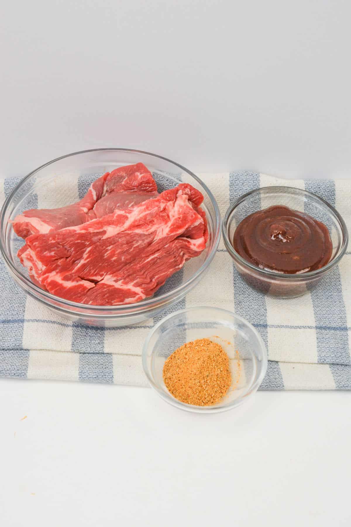 Three glass bowls on a striped towel: one with raw beef slices  one with brown barbecue sauce, and one with a dry seasoning mix.