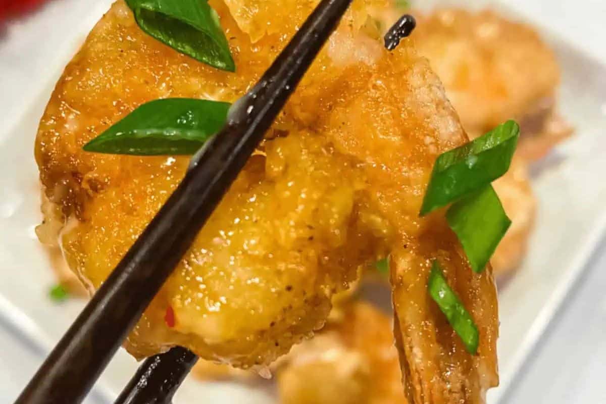 Chopsticks delicately grasp a restaurant-quality crispy fried bang bang shrimp, perfectly garnished with sliced green onions.