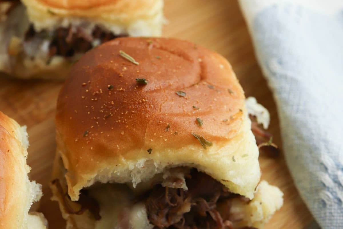 Close-up of a slider sandwich with shredded meat and melted cheese on a soft bun, garnished with herbs—an easy dinner favorite from popular slow cooker recipes—placed on a wooden surface near a blue and white cloth.