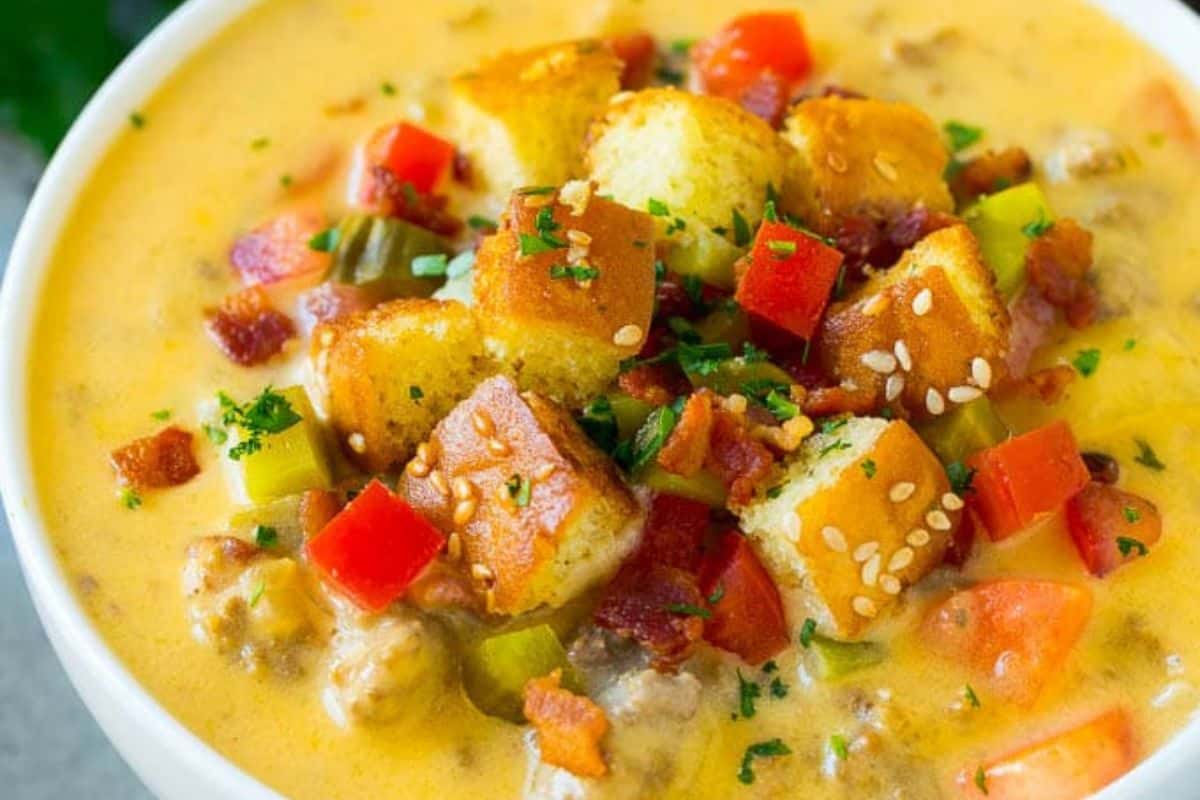 Close-up of a white bowl of cheeseburger soup, topped with tomatoes, bacon, croutons, and sesame seeds.