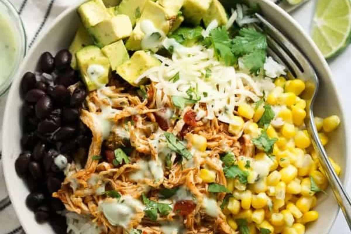 A bowl with shredded chicken, black beans, corn, diced avocado, shredded cheese, rice, and cilantro, drizzled with a creamy dressing—perfect for spicing up your dinner routine. Served with a fork and a lime wedge on the side.