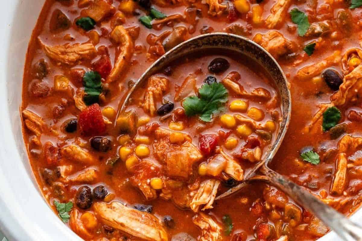 A ladle scoops up chunky chicken tortilla soup with black beans, corn, tomatoes, and cilantro from a white bowl—one of the best easy dinner recipes for a comforting meal.