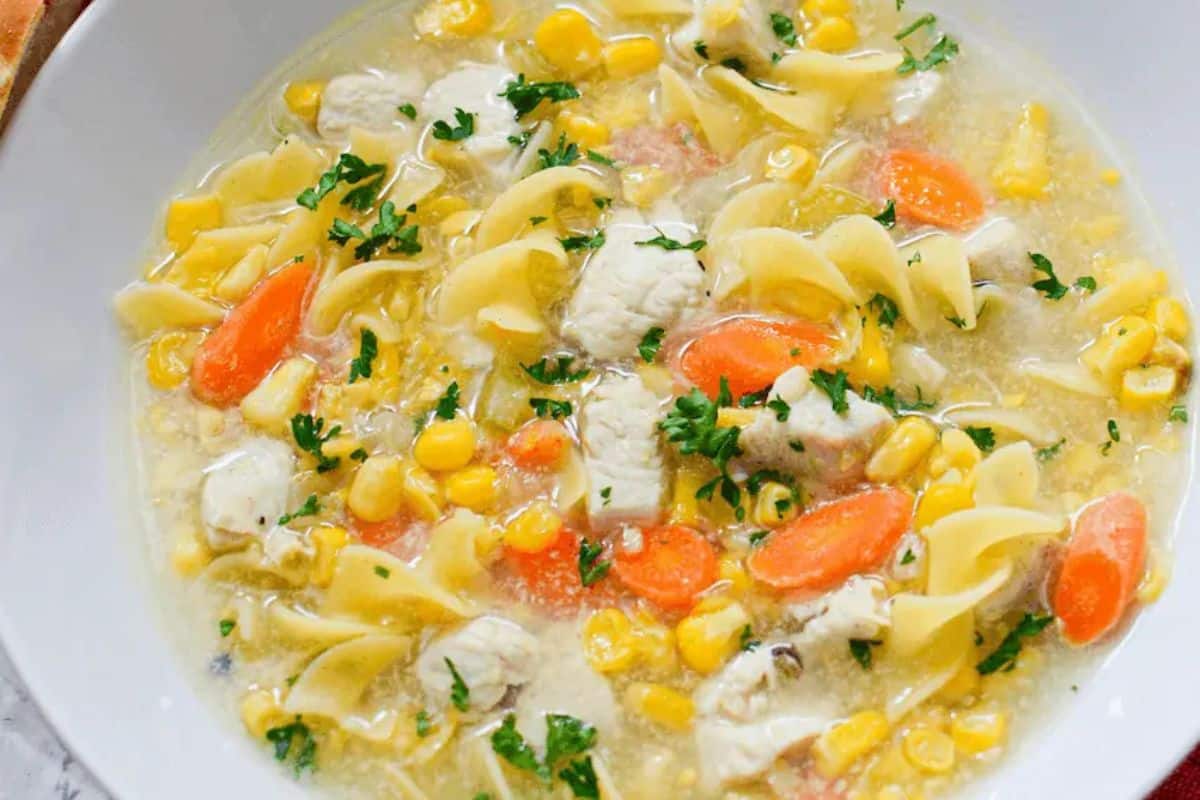 Close-up of a white bowl of chicken corn noodle soup with noodles, carrots, corn, and chicken.