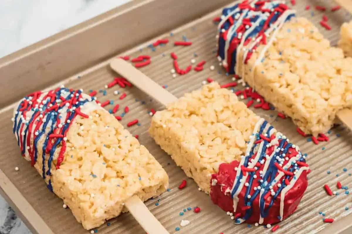 Irresistible Rice Krispie treats on sticks, decorated with red, white, and blue icing and sprinkles, showcased on a baking sheet. Perfect for any snack game moment!.