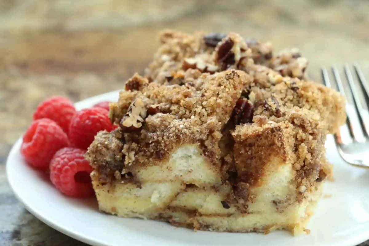 A slice of a cinnamon roll french toast bake with a streusel topping on a white plate, perfect for breakfast or brunch, accompanied by a few raspberries and a fork.