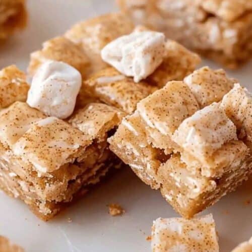 Close-up of cereal bars topped with marshmallows and dusted with cinnamon on a white surface, these delicious treats bring comfort in every bite.