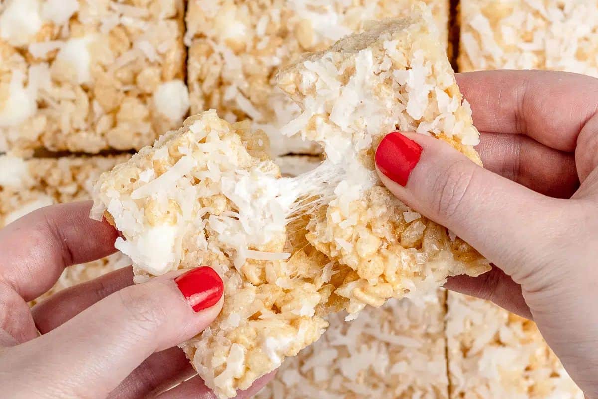 Hands adorned with red nail polish pull apart an irresistible rice crispy treat, revealing gooey marshmallow and coconut flakes. Against a backdrop of similar treats, this scene elevates your snack game to new heights.