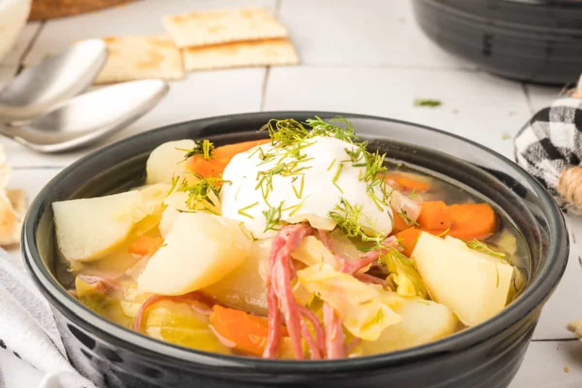 A bowl of soup with potatoes, carrots, and tender corned beef, topped with sour cream and dill, sits gracefully on a table alongside crackers and spoons. This modern take on traditional recipes brings warmth and comfort to any meal.
