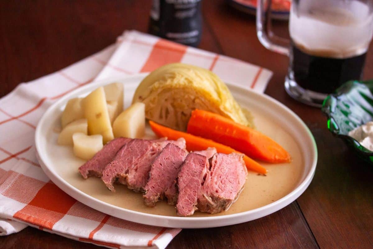 Celebrate the season with a plate of sliced corned beef, carrots, boiled potatoes, and cabbage wedge on a tablecloth. A glass of dark beverage adds the finishing touch to this classic cabbage recipe setting.