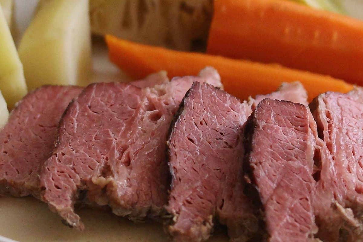 Traditional slices of cooked corned beef rest elegantly on a plate, with vibrant carrots and classic cabbage recipes complementing the background.