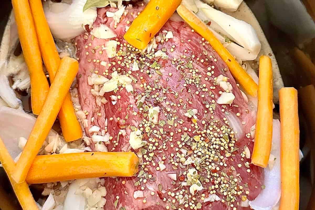 A raw beef roast in a crockpot with chopped carrots, onions, garlic, and sprinkled spices brings a modern corned beef recipe to life.