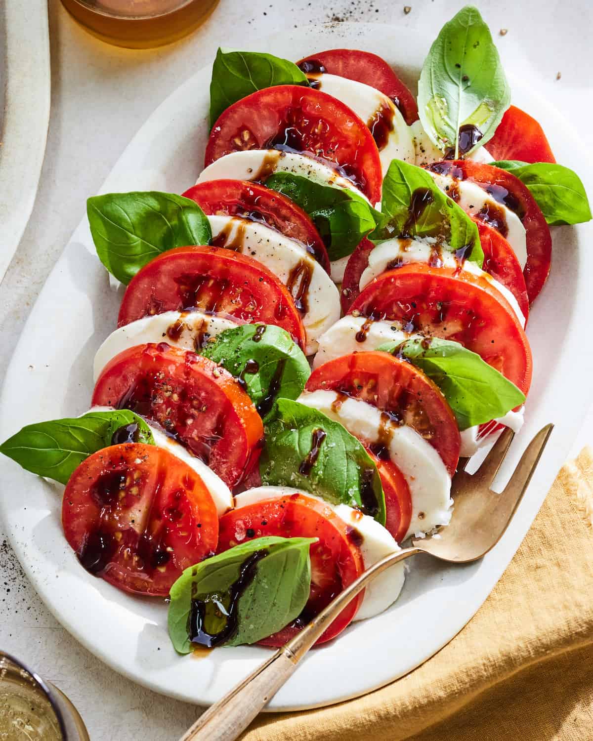 A caprese salad on a white platter, featuring sliced tomatoes, mozzarella, basil leaves, and balsamic glaze—perfect for potluck dishes. A fork and beige napkin sit beside this foolproof recipe that's sure to impress.
