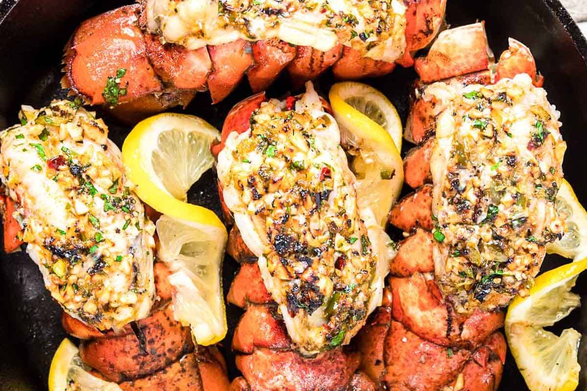 Close-up of cooked lobster tails with a buttery herb topping and lemon slices.