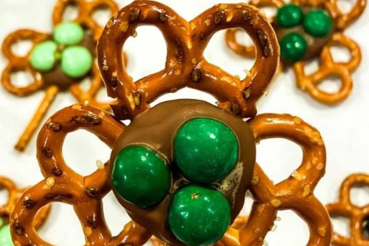 A close-up of shamrock-shaped treats made from pretzels, melted chocolate, and green candy-coated chocolates, arranged on a white surface. The pretzels form the leaves of the shamrock, with the chocolate acting as a base to hold the green candies in place.